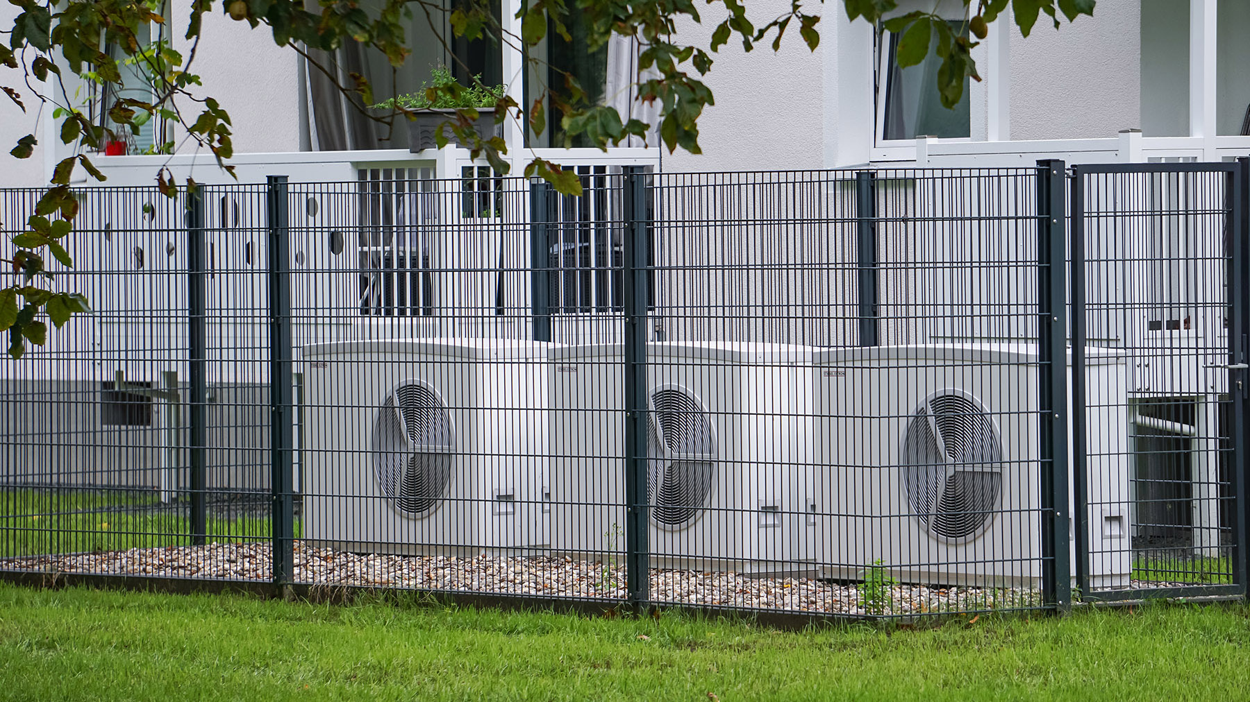 Drei Wärmepumpen stehen eingezäunt vor einer Wohnsiedlung in Köln. 