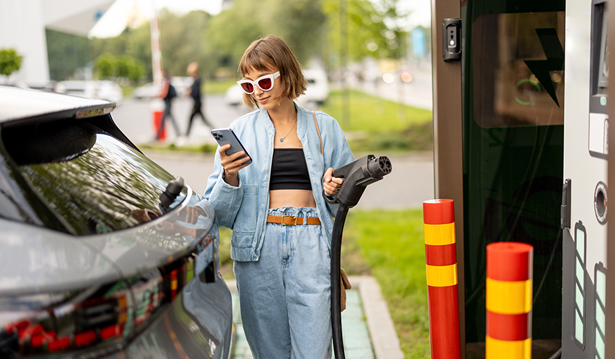 Junge Frau checkt ihr Handy während sie das Ladekabel einer Schnellladestation in der Hand hält.