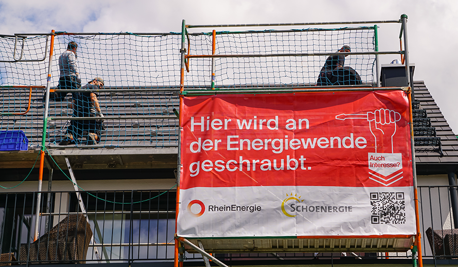 Vor einem Hausdach hängt ein Baustellenbanner mit dem Schriftzug:; Hier wird an der Energiewende geschraubt.